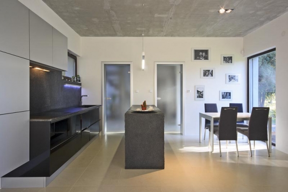V minimalisticky pojatém interiéru kladli majitelé domu důraz na čisté tvary a kvalitní materiály – pohledový beton, sklo, lakované MDF desky s vysokým leskem. Kuchyňská linka má pracovní desku, obložení stěny a bočnice z tmavé žuly. Dominantu obývacího pokoje představují nadčasová funkcionalistická křesla Barcelona (design Mies van der Rohe).