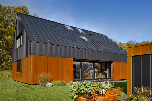 Kompaktní fasáda jednoduchého domu je členěna vertikálními falcovanými spoji cortenového a černého plechu. Barevný kontrast doplňují prosklené plochy.